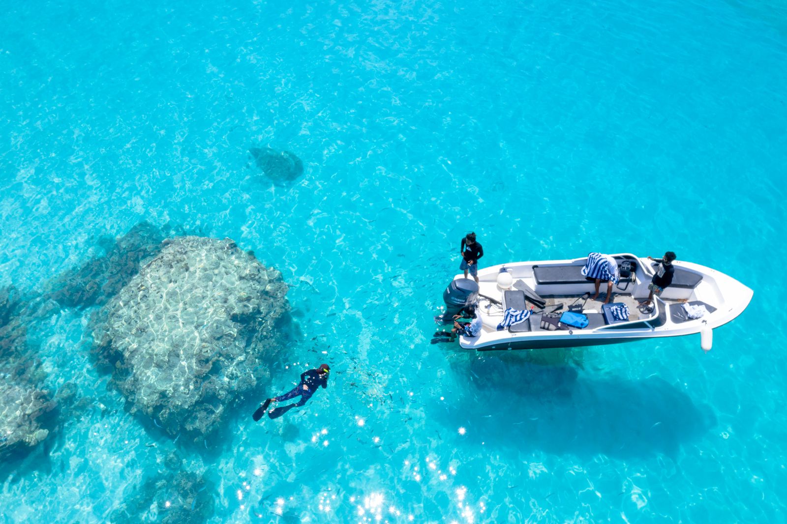透明な浅い海に停まった小型ボートのそばで、ウェットスーツを着た人がシュノーケリングをしている様子を上空から見た写真（イメージ写真・沖縄ではありません）